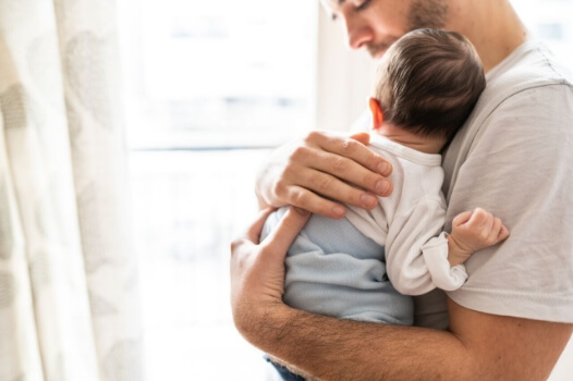 Ein junger Mann hält liebevoll ein Baby, das seinen Kopf auf seine Schulter legt – Symbol für sanfte Pflege mit Tannosynt bei Windeldermatitis.