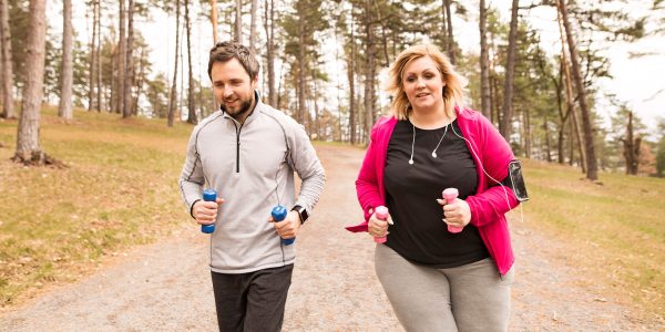 Im Bildhintergrund sind ein Wald und ein Feldweg zu erkennen, auf dem zwei Personen erscheinen. Ein Junge mit zwei kleinen Trainingsgewichten in seinen Händen. Er trägt ein graues Sweatshirt und eine schwarze Sporthose. Zu seiner Rechten ein Mädchen, das ein offenes rosa Sweatshirt und eine graue Jogginghose trägt. In seinen Händen trägt er außerdem ein Paar kleine Sportgewichte, einige Kopfhörer, die an eine Hülle für ein Arm-Handy angeschlossen sind